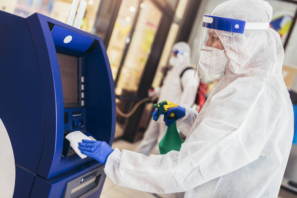 Professional workers in hazmat suits disinfecting ATM machine. - Photo, Image