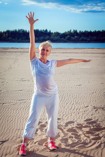 Femme aux cheveux blonds courts en survêtement blanc sur une plage de sable fait des exercices
 - Photo, image