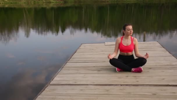 Dziewczyna ćwiczy technikę jogi. Siedzi na molo w lotosowej pozycji nad jeziorem. - Materiał filmowy, wideo