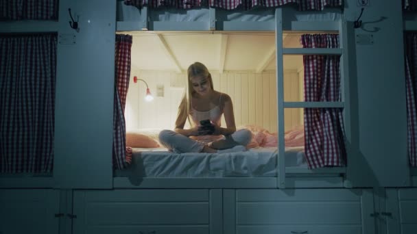Mujer atractiva joven sentado en la cama en la habitación del albergue y el uso de teléfono inteligente por la noche - Imágenes, Vídeo