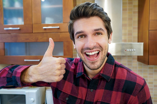 młody przystojny szczęśliwy człowiek z duży uśmiech pokazując kciuk w górę znak w kuchnia mając na sobie koszulę - Zdjęcie, obraz