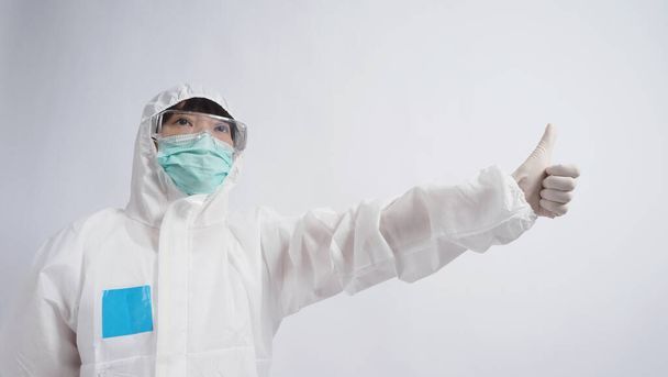 Imágenes de gran angular de la mujer asiática médico en traje PPE o equipo de protección personal y gafas médicas, máscara, guantes y mostrando el pulgar hacia arriba o como simbólico que representan un momento feliz con baja pandemia
  - Foto, imagen