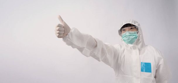 Laajakulmainen kuva aasialaisesta naislääkäristä PPE-puvussa tai henkilönsuojaimissa ja lääketieteellisissä suojalaseissa, naamiossa, käsineissä ja peukalon pystyssä tai symbolisena, jotka edustavat onnellista hetkeä, jossa pandemia on alhainen
  - Valokuva, kuva