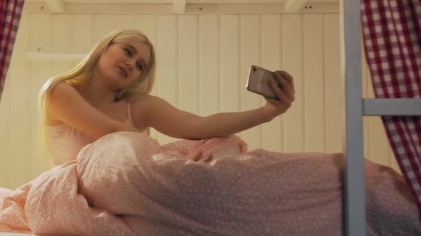 Primer plano de la mujer atractiva joven sentada en la cama en la habitación del albergue y el uso de teléfono inteligente que tiene chat de vídeo - Imágenes, Vídeo