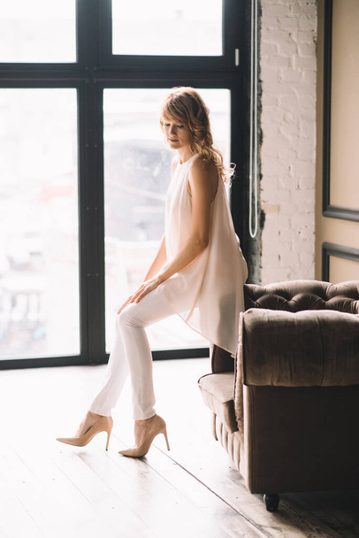 Hermosa joven mujer rubia delgada con el pelo rizado en pantalones blancos, una blusa y zapatos en un estudio loft cerca de la ventana. Enfoque selectivo suave
. - Foto, imagen