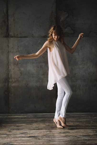 Schöne junge schlanke blonde Frau mit lockigem Haar in weißen Hosen, Bluse und Schuhen in einem Loft-Studio tanzt. Weiche selektive Fokussierung. - Foto, Bild