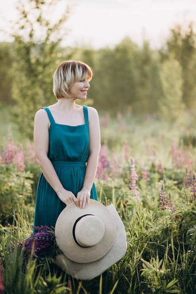 Beautiful romantic girl blonde in a dress in a field of purple flowers of lupins at dawn holds a hat in her hands looks into the distance and smiles. Soft selective focus. - Fotó, kép