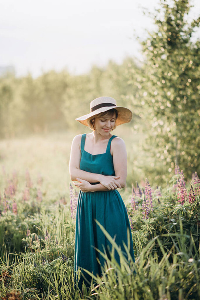 Şafak vakti mor lupin çiçekleriyle dolu bir tarlada elbisesi ve şapkası olan güzel romantik bir kız. Yumuşak seçici odak. - Fotoğraf, Görsel