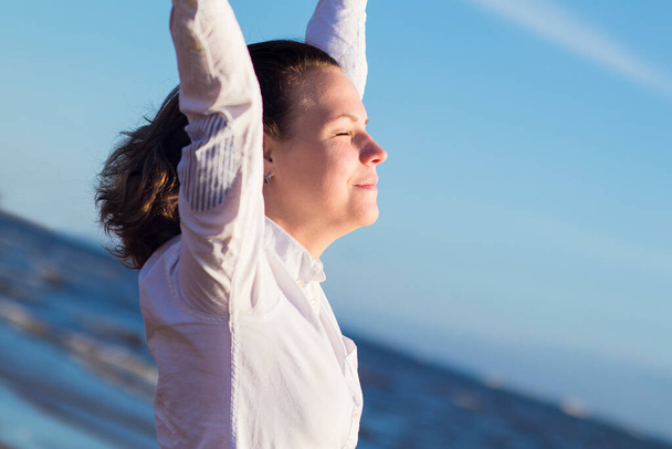 Happy positive beautiful girl, young woman in white shirt enjoying vacation on sea, beach, stretch, hold her arms, hands up with eyes closed. Good mood, relaxation, happiness concept - Foto, Bild