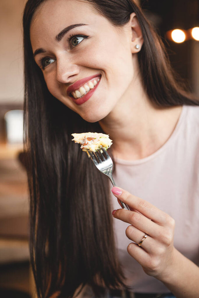 woman with exquisite smile and beautiful eyes having breakfast with her friend in a cozy cafe. She eats a fork a piece of cake and drinks tea sitting in a chair at a table - Fotografie, Obrázek