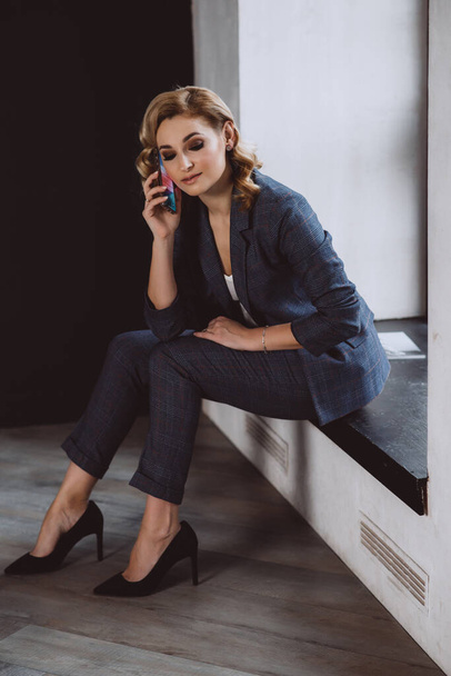 Portrait of a confident beautiful business woman in a suit sitting on the window talking on the phone. The concept of gender equality. Strong independent woman. Soft selective focus. - Fotografie, Obrázek