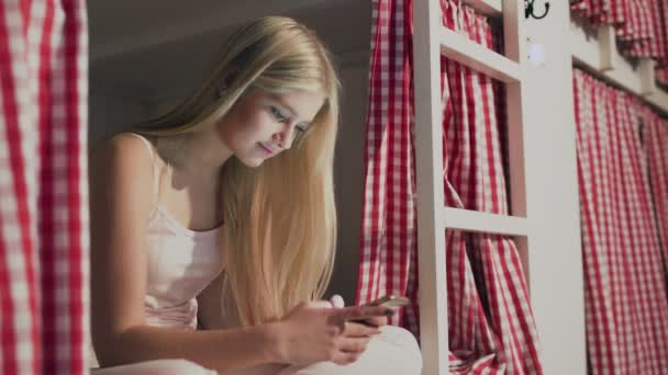 Jonge aantrekkelijke vrouw zitten op het bed in het hostel kamer en met behulp van smartphone - Video
