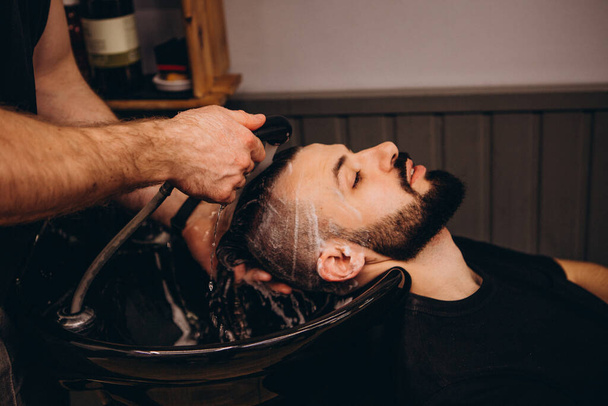 Handsome bearded man is having his hair washed by hairdresser at the barbershop. customer service in the beauty salon, haircut and hair washing concept - Valokuva, kuva