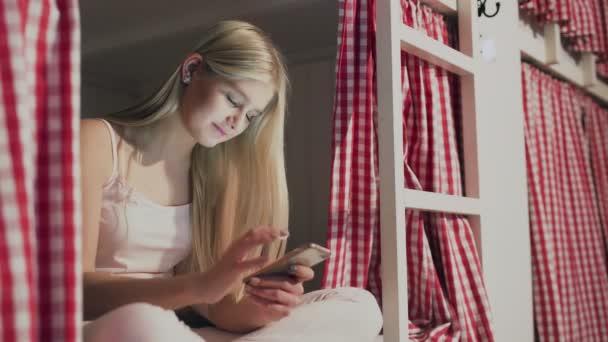 Fiatal vonzó nő ül az ágyon a hostel szobában, és használja okostelefon - Felvétel, videó
