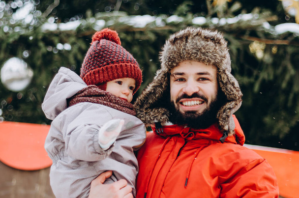 Young stylish bearded Father and baby Son On Winter Vacation near Christmas Tree. Father giving son ride on back in park. Happy, joyful family. - Zdjęcie, obraz