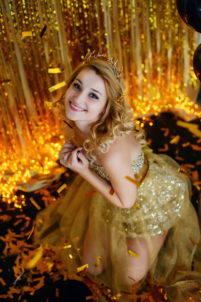 Όμορφη νεαρή κοπέλα μοντέλο ξανθά χαμόγελα σε ένα κομψό χρυσό φόρεμα με κέρατα στεφάνης φόντο λωτού κορδέλες με γιρλάντες κάθεται στο πάτωμα και με ένα χρυσό metophan καραμέλα. - Φωτογραφία, εικόνα