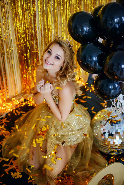Güzel genç kız model sarışın zarif altın bir elbiseyle gülümsüyor. Arka planda nilüfer çiçekli kurdeleler ve altın bir şeker metofanı var.. - Fotoğraf, Görsel