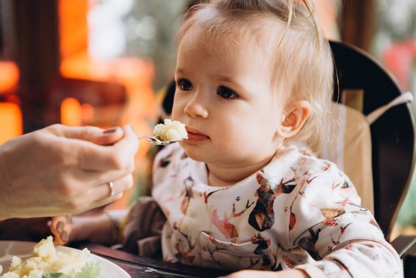 Anne bebeğini kaşıkla besler ve küçük bir masada oturur. Yaramaz çocuk ağlıyor ve yemek yemek ve ağlamak istemiyor. - Fotoğraf, Görsel