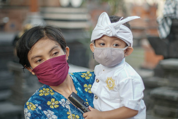 Balijska matka i dziecko noszą balijskie tradycyjne ubrania podczas pandemii korony lub covid-19. Obaj używają masek, by chronić się przed atakami wirusów.. - Zdjęcie, obraz