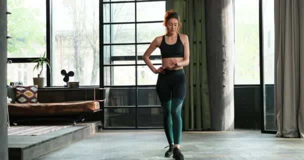 Young Caucasian sporty woman standing in living room and warming up her legs. Female training her body on quarantine self-isolation. Pretty girl training and working out at home. - Filmmaterial, Video