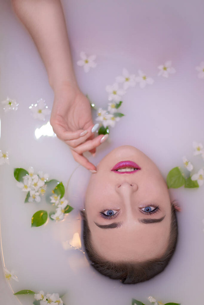 A charming girl with stylish makeup and beautiful blue eyes is taking a milk bath with a pink hue strewn with Apple blossoms and leaves. Touches the sweet face with gentle hands. - Photo, image