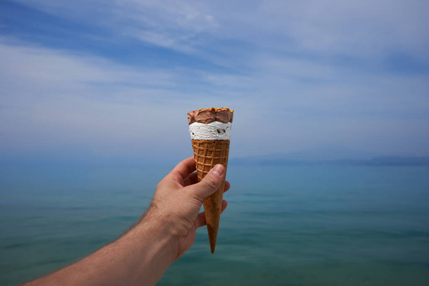 A hand with cone of bited melting ice cream gelato with a background of blue sky and lake on bright summer day - Photo, Image