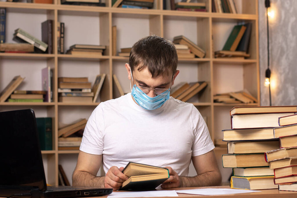 Jongeman met een medisch masker zit aan een tafel met laptops en leest een boek. Moeilijkheden met online leren in quarantaine. kantoorwerk op afstand. kast op de achtergrond. - Foto, afbeelding