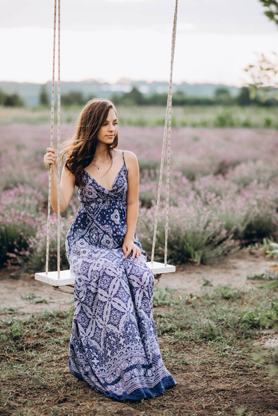 красива дівчина в довгій сукні сидить на гойдалці в лавандовому полі на заході сонця. М'який фокус
. - Фото, зображення