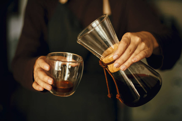 Professionelle Barista in schwarzer Uniform, die Tropfkaffee zubereitet. Person gießt frischen aromatischen Kaffee aus Glaskolben in Tasse, Hände in Großaufnahme. Alternative Kaffeezubereitung, v60. - Foto, Bild