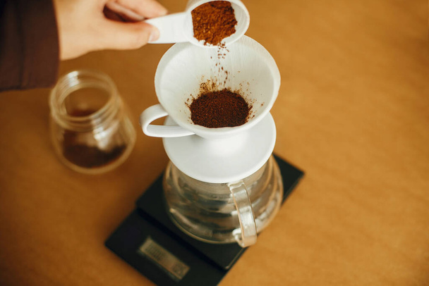 Ręce wlewające uziemioną kawę do filtra zalać szklanym czajnikiem w skali. Przygotowanie do alternatywnego zaparzania kawy v60. Osoba trzymająca łyżkę z mieloną kawą na brązowym tle - Zdjęcie, obraz