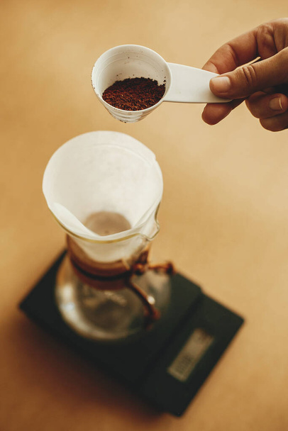 Kädet kaatamalla maadoitettu kahvi suodatin kaada päälle lasi vedenkeitin mittakaavassa. Valmistautuminen vaihtoehtoiseen kahvinkeittoon v60. Henkilö jolla lusikka jauhaa kahvia ruskealla taustalla
 - Valokuva, kuva