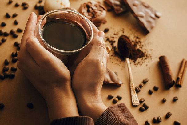 Ruce držící skleněný šálek s čerstvou kávou a praženými fazolemi, mletá káva na lžíci, makarony, čokoláda, skořice na hnědém pozadí. Káva náladový obrázek - Fotografie, Obrázek