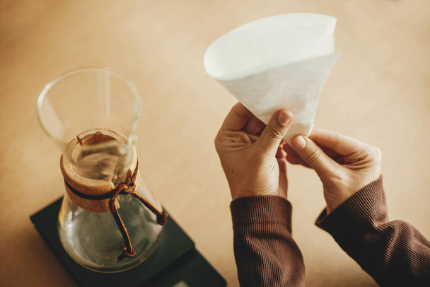 Руки складывают бумажный фильтр для заливки поверх и стеклянный чайник на шкале на коричневом фоне. Подготовка к альтернативному варке кофе v60. Сложите фильтр кофе
 - Фото, изображение