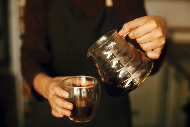Людина виливає свіжу ароматну каву зі скляного чайника в чашку. Альтернативне пивоварне виробництво кави, v60. Професійна бариста в чорній формі робить крапельницю кавою
 - Фото, зображення