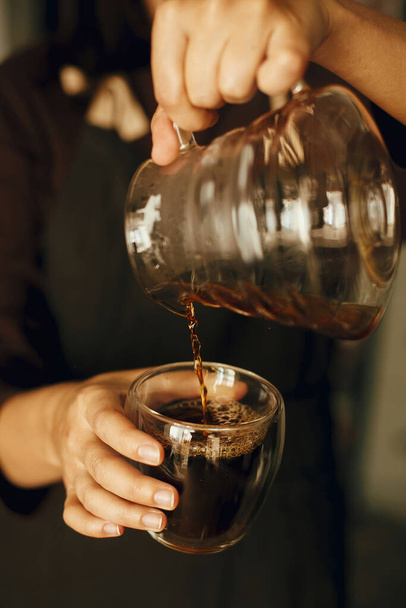 Person pouring fresh aromatic coffee from glass kettle in cup. Alternative coffee brewing, v60. Professional barista in black uniform making drip coffee - Foto, Bild