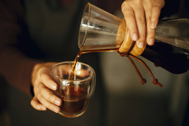 Professional female barista in black uniform making drip coffee. Person pouring fresh aromatic coffee from glass flask in cup, hands close up. Alternative coffee brewing, v60. - Foto, afbeelding