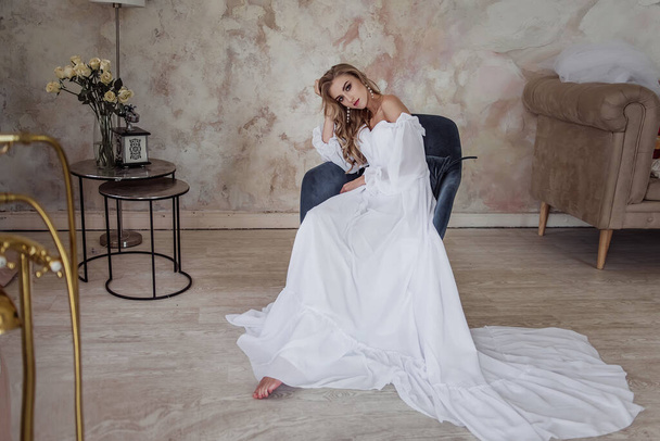 A beautiful girl with big, expressive eyes, long legs and blond hair in a white dress sitting on a chair in a stylish loft - a modern image of the bride. selective back and front focus, art noise - Фото, изображение
