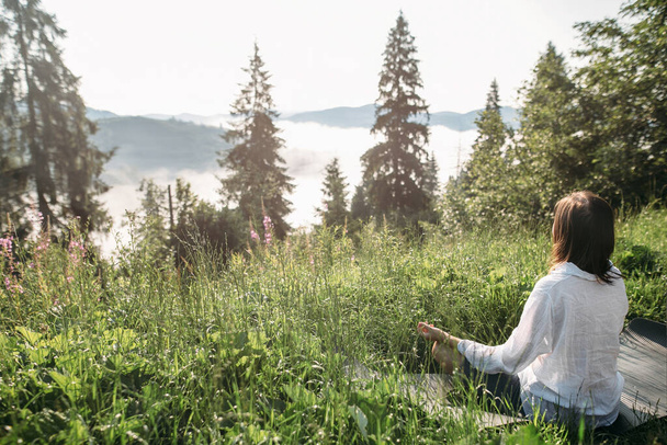 Young woman practicing yoga and sitting in lotus position on top of mountain among grass in sunny morning light. Fit girl meditating. Calm tranquil moment, connection with nature - Photo, Image