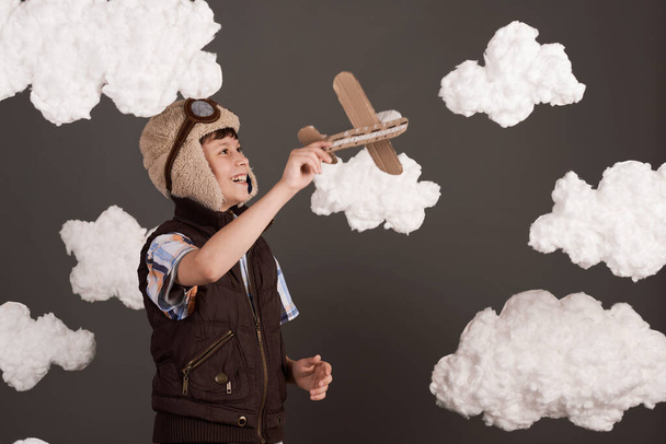 een jongen speelt met een kartonnen vliegtuig en droomt ervan piloot te worden, gekleed in een retro jasje en helm met bril, wollen wolken, grijze achtergrond, bruin getint - Foto, afbeelding
