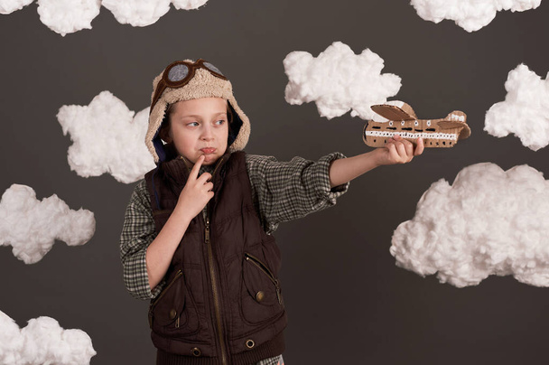 a girl plays with a cardboard airplane and dreams of becoming a pilot, dressed in a retro style jacket and helmet with glasses, clouds of cotton wool, gray background, tinted in brown - Photo, Image