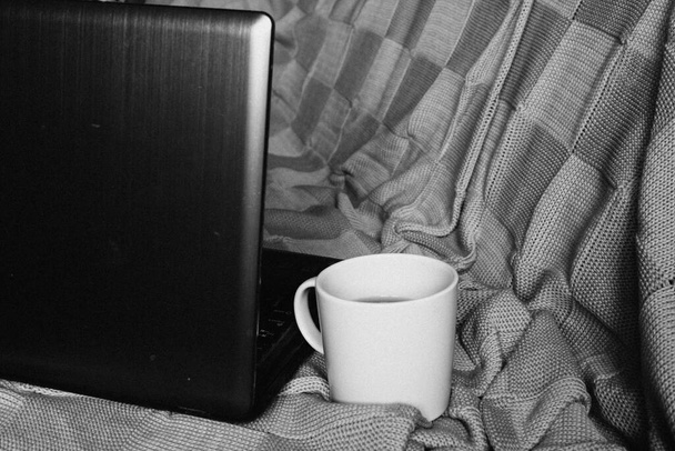 Styl życia wolnego strzelca. Praca zdalna z domu podczas samoizolacji. Laptop i filiżanka herbaty z cytryną na wygodnym kocu z dzianiny na łóżku w czerni i bieli - Zdjęcie, obraz
