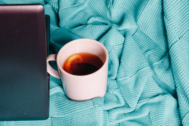 Der Lebensstil eines Freiberuflers. Fernarbeit von zu Hause aus während der Isolation. Laptop und eine Tasse Tee mit Zitrone auf einer gemütlichen gestrickten blauen (türkisfarbenen) Decke auf dem Bett mit Kopierraum. - Foto, Bild
