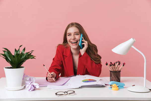 Image of a happy young woman sit at the table indoors isolated over pink wall background talking by mobile phone writing notes. - Фото, зображення