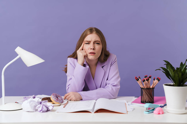 Photo of a serious concentrated young woman sit at the table indoors isolated over purple wall background. - 写真・画像