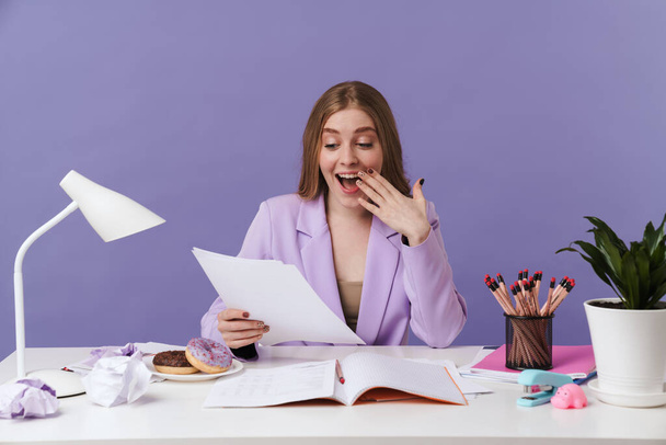 Image of a positive surprised young woman sit at the table indoors isolated over purple wall background holding documents. - Foto, imagen
