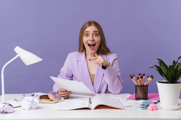 Image of a shocked excited young woman sit at the table indoors isolated over purple wall background holding documents. - Foto, Imagem