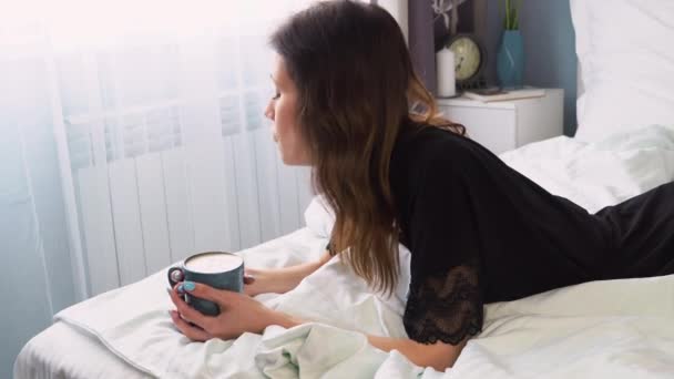 Side view woman in black bathrobe lying in bed and have cup of coffee, enjoying coffee at morning time - Imágenes, Vídeo