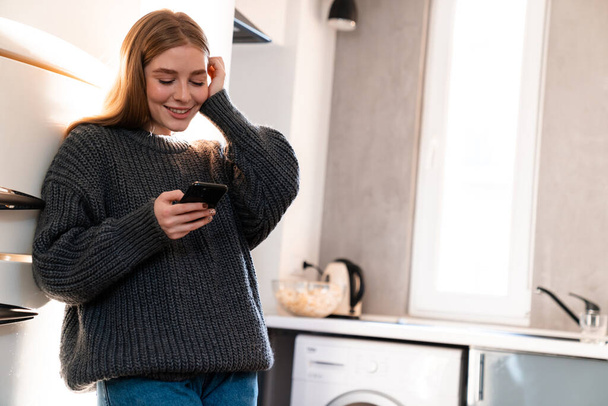 Image of a pleased positive young woman indoors at home using mobile phone. - Foto, Imagen