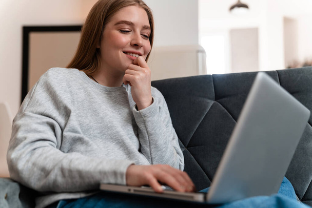 Photo of a happy positive beautiful young woman indoors at home on sofa using laptop computer. - Valokuva, kuva