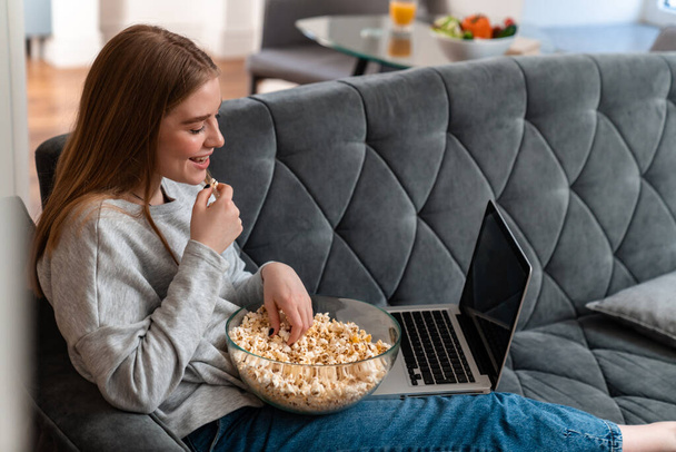 写真の正楽観的な美しい若い女性屋内で自宅でソファの上にラップトップコンピュータの時計映画を使用してポップコーンを食べる. - 写真・画像
